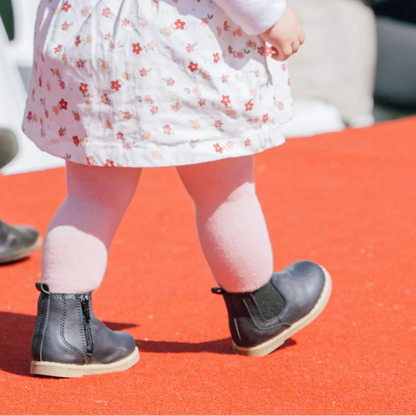 A toddler walks down a red carpet at an outdoor event, wearing a white floral dress, pink merino wool tights, and black boots