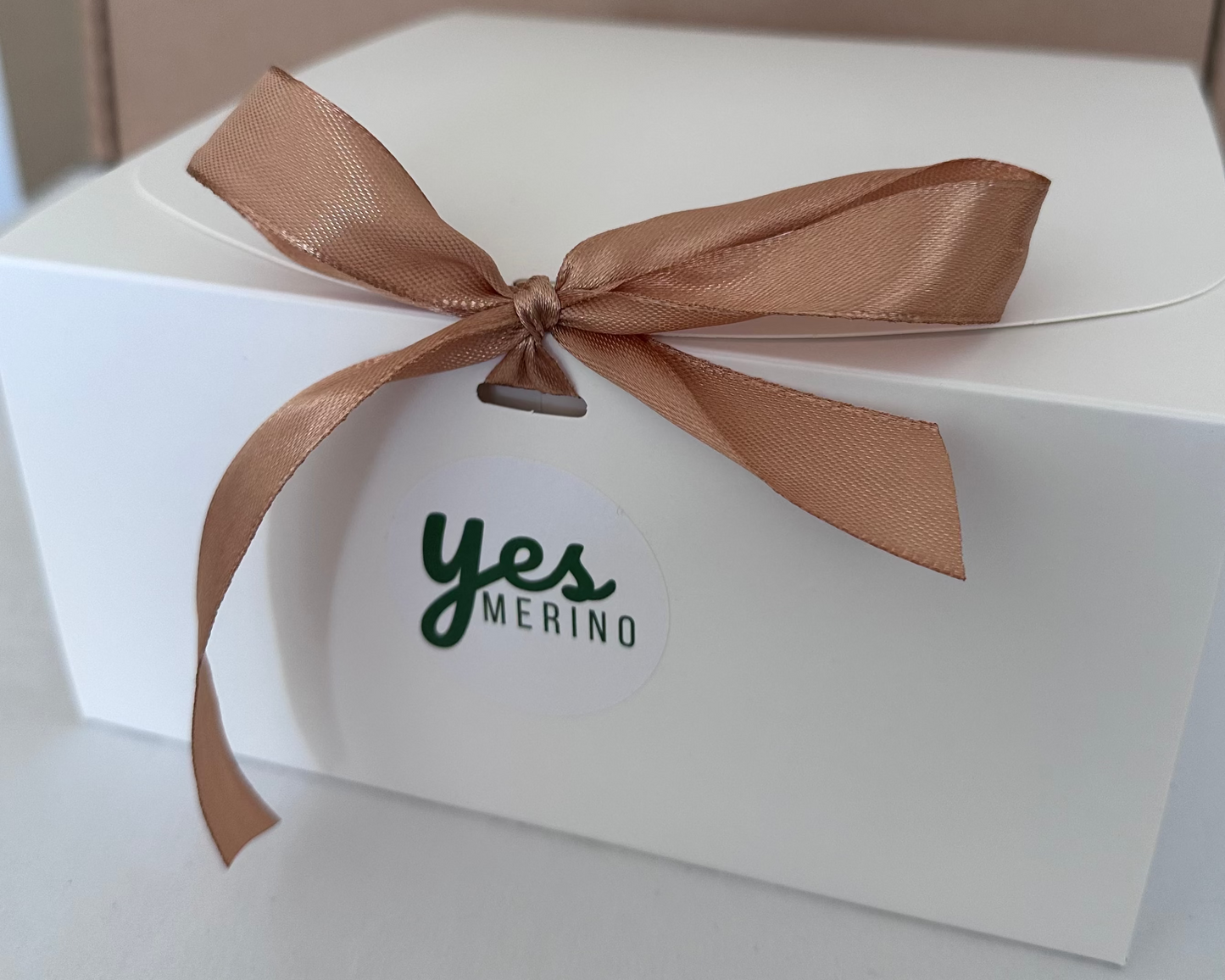 A white square gift box tied with a metallic bronze satin ribbon bow and a small hanging tag that reads 'yes MERINO', set against a neutral background.