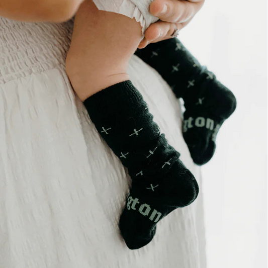 Mother holding a  baby wearing a pair of dark green merino wool socks with light green crosses and Lamington knitted across the top of the foot.