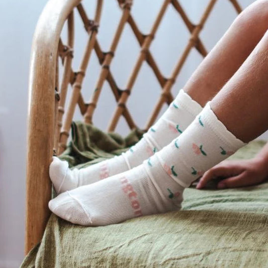 A child sits on a rattan chair wearing white merino wool crew socks with small flower prints and the word 'Lamington' visible on one sock.